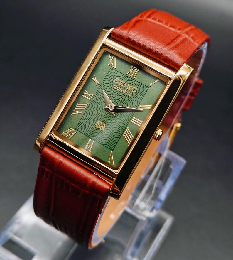 Puede incluir: Un reloj Seiko Quartz rectangular con caja dorada y esfera verde con n&uacute;meros romanos dorados. El reloj tiene una correa de cuero roja. El reloj se exhibe sobre un soporte transparente sobre un fondo oscuro.