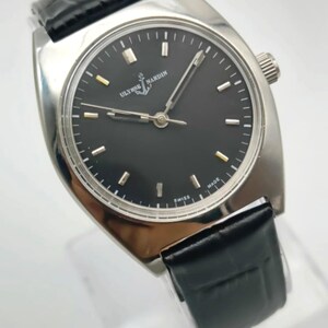 May include: A silver-toned Ulysse Nardin wristwatch with a black face and a black leather band. The watch face has silver markers and hands, with the brand name and anchor logo visible. The watch is marked "SWISS MADE".