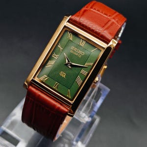Puede incluir: Un reloj Seiko Quartz rectangular dorado con esfera verde y n&uacute;meros romanos. El reloj tiene una correa de cuero marr&oacute;n. La esfera del reloj presenta un patr&oacute;n texturizado y el logotipo de Seiko Quartz.