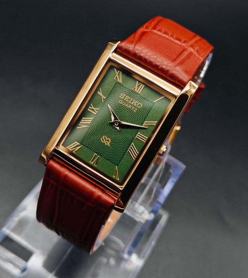 Puede incluir: Un reloj Seiko Quartz rectangular dorado con esfera verde y n&uacute;meros romanos. El reloj tiene una correa de cuero marr&oacute;n. La esfera del reloj presenta un patr&oacute;n texturizado y el logotipo de Seiko Quartz.