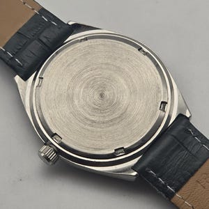 K&ouml;nnte beinhalten: Nahaufnahme der R&uuml;ckseite einer silbernen Armbanduhr mit schwarzem Lederarmband. Das Zifferblatt der Uhr ist in geb&uuml;rstetem Silber gehalten und hat ein kreisf&ouml;rmiges Design. Das schwarze Lederarmband ist mit wei&szlig;em Faden gen&auml;ht.
