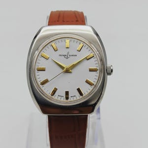 K&ouml;nnte beinhalten: Eine Vintage-Armbanduhr von Ulysse Nardin mit silberfarbenem Geh&auml;use und wei&szlig;em Zifferblatt. Die Uhr hat goldfarbene Zeiger und Markierungen sowie ein braunes Lederarmband. Die Uhr hat einen Durchmesser von ca. 3,8 cm.