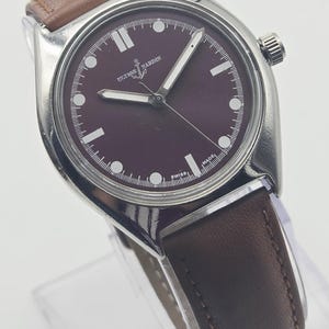 May include: A silver-toned wristwatch with a burgundy face and brown leather band. The watch face features white hour markers and hands, with the brand name "Ulysse Nardin" visible. The watch is displayed on a clear stand.