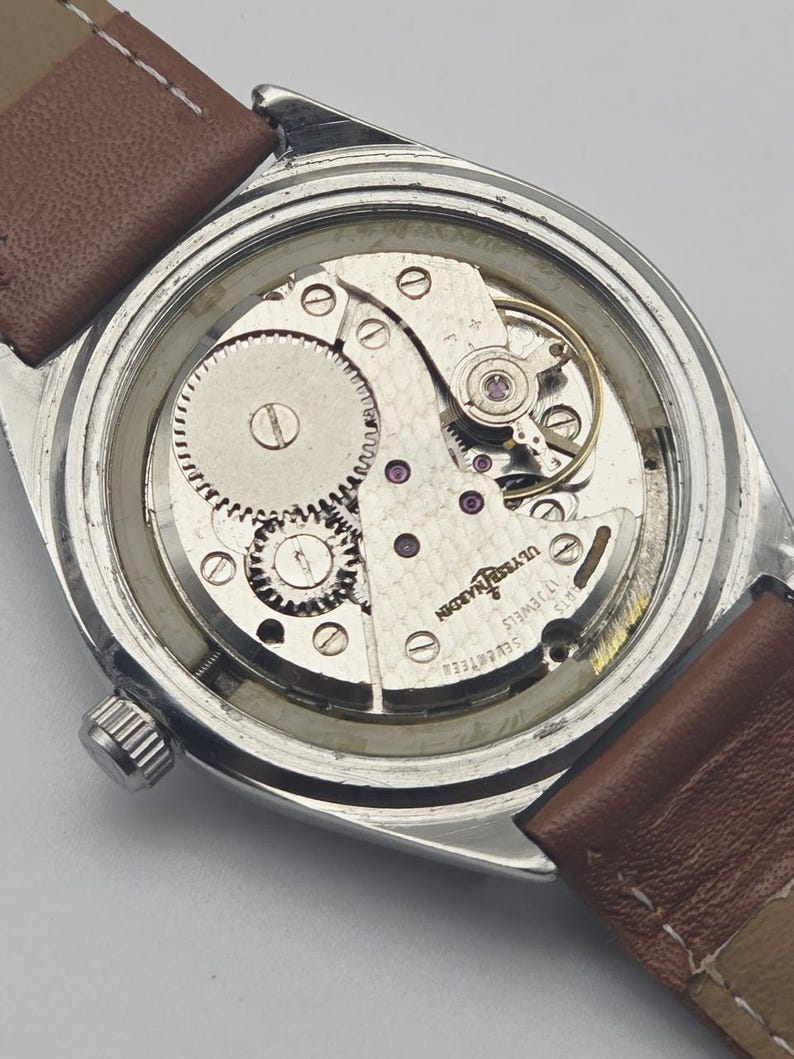 May include: A vintage silver-toned wristwatch with a brown leather strap. The watch's back is open, revealing the intricate mechanical movement, including gears, screws, and the inscription "ULYSSE NARDIN 17 JEWELS".