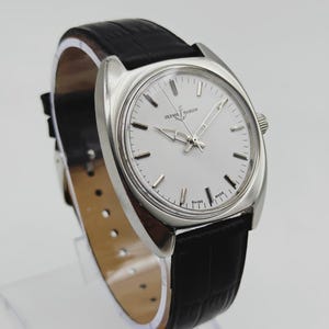 May include: A silver Ulysse Nardin wristwatch with a white face and a black leather strap. The watch features a round dial with silver hands and markers. The strap is textured and has a buckle. The watch is displayed on a clear stand.