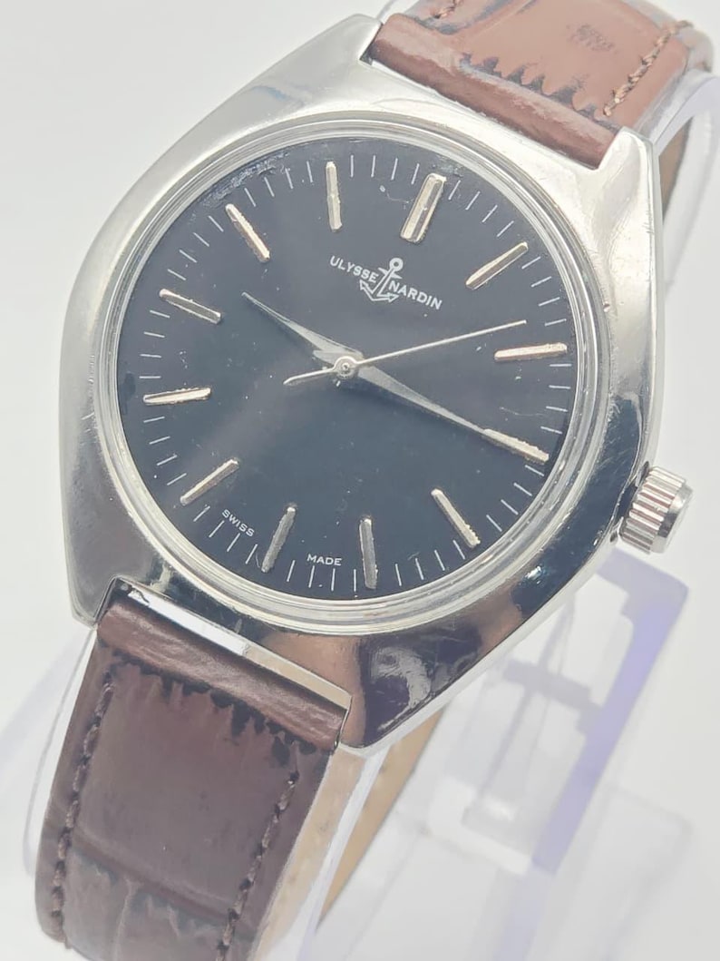 May include: A vintage Ulysse Nardin wristwatch with a black dial and silver-tone hands and markers. The watch features a stainless steel case and a brown leather strap. The watch face displays the brand name and the words "Swiss Made".
