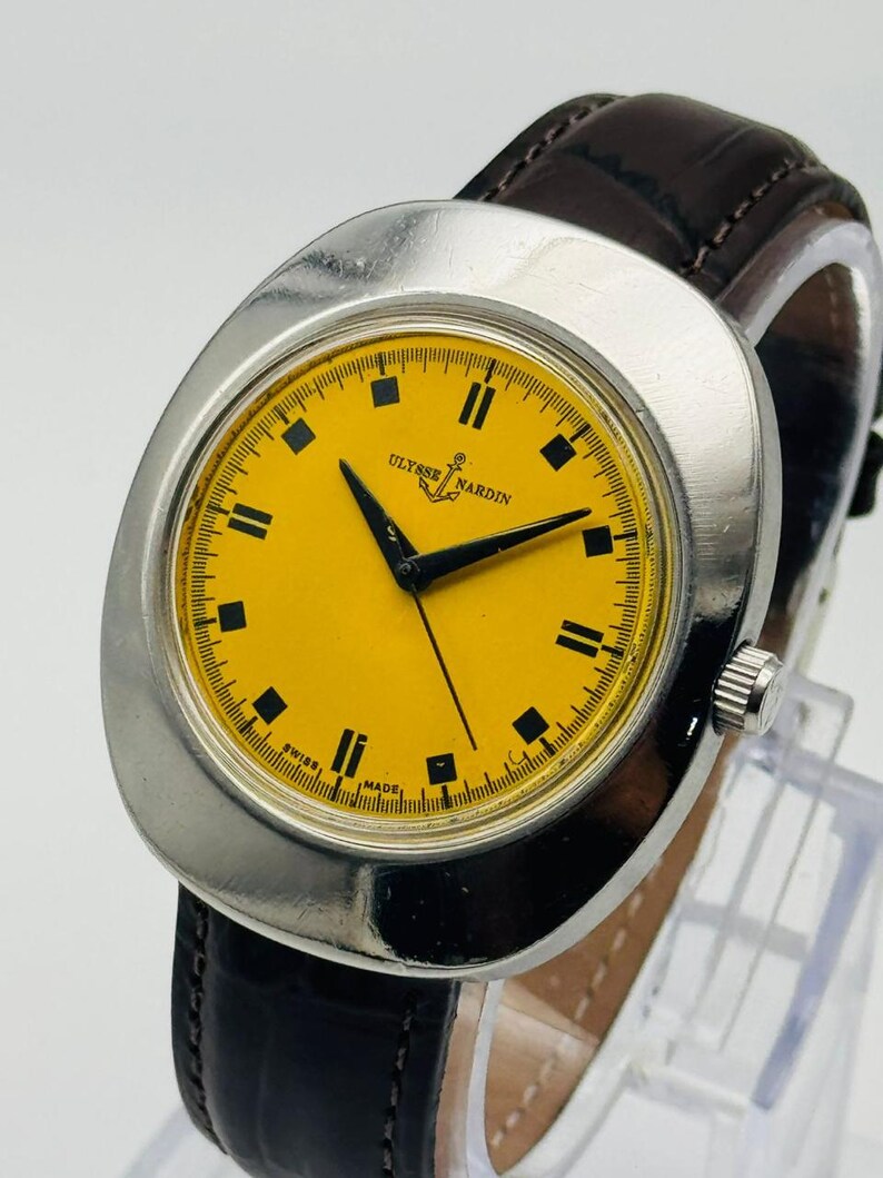 May include: A vintage Ulysse Nardin wristwatch with a silver-tone case and a bright yellow dial. The watch features black hour markers and hands, and a brown leather strap. The watch face has the text "Ulysse Nardin" and "Swiss Made".