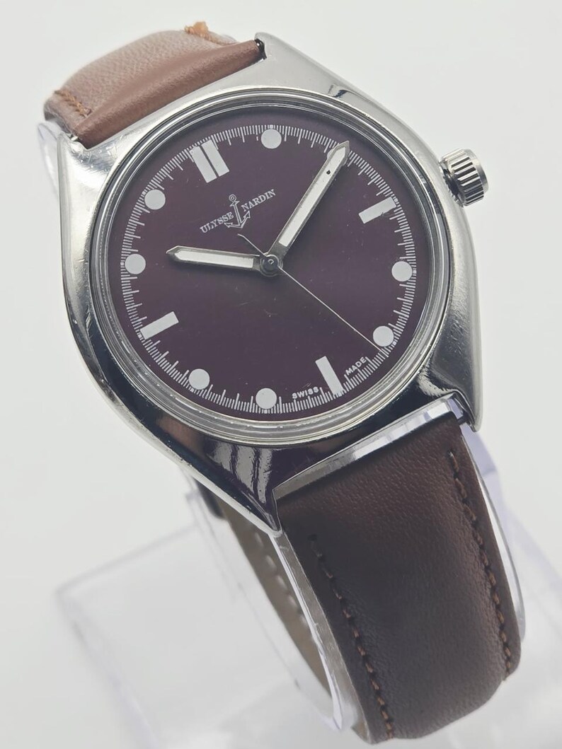 May include: A silver-toned wristwatch with a burgundy face and brown leather band. The watch features white hour markers and hands, with the brand name "Ulysse Nardin" visible on the face. The watch is displayed on a clear stand.