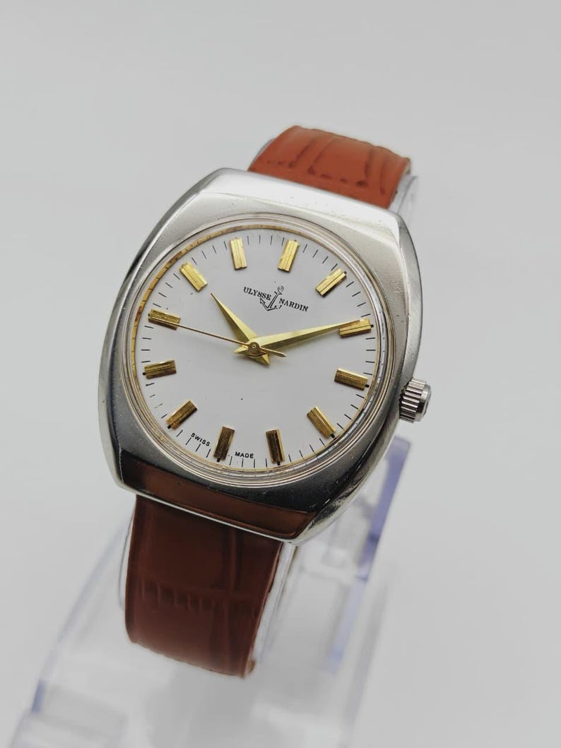 K&ouml;nnte beinhalten: Eine Vintage-Armbanduhr von Ulysse Nardin mit silberfarbenem Geh&auml;use und wei&szlig;em Zifferblatt. Die Uhr hat goldfarbene Zeiger und Stundenmarkierungen sowie ein braunes Lederarmband. Die Uhr ist mit "Swiss Made" gekennzeichnet.