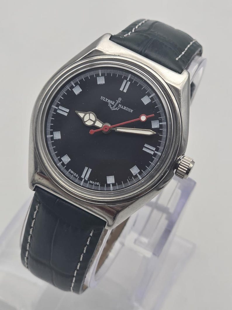 K&ouml;nnte beinhalten: Eine Ulysse Nardin Armbanduhr mit schwarzem Zifferblatt und schwarzem Lederarmband. Die Uhr hat ein silbernes Geh&auml;use, wei&szlig;e Stundenmarkierungen und einen roten Sekundenzeiger. Die Uhr wird auf einem transparenten St&auml;nder pr&auml;sentiert.