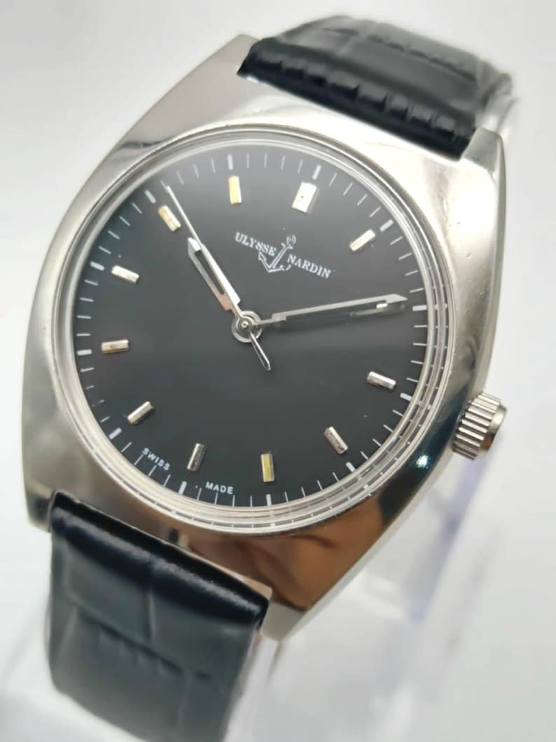 May include: A silver-toned Ulysse Nardin watch with a black face and a black leather band. The watch face has silver and gold-toned markers and hands. The band has a crocodile pattern. The watch is marked "SWISS MADE".