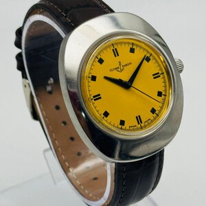 May include: A vintage Ulysse Nardin wristwatch with a yellow face and black hands. The watch has a silver-colored case and a dark brown leather band. The watch face displays the brand name and is marked "Swiss Made".