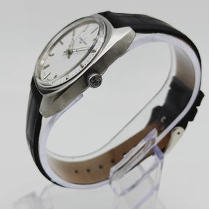 May include: A silver wristwatch with a white face and a black leather strap. The watch features a round face with silver hands and markers. The black strap is fastened with a silver buckle. The watch is displayed on a clear stand.