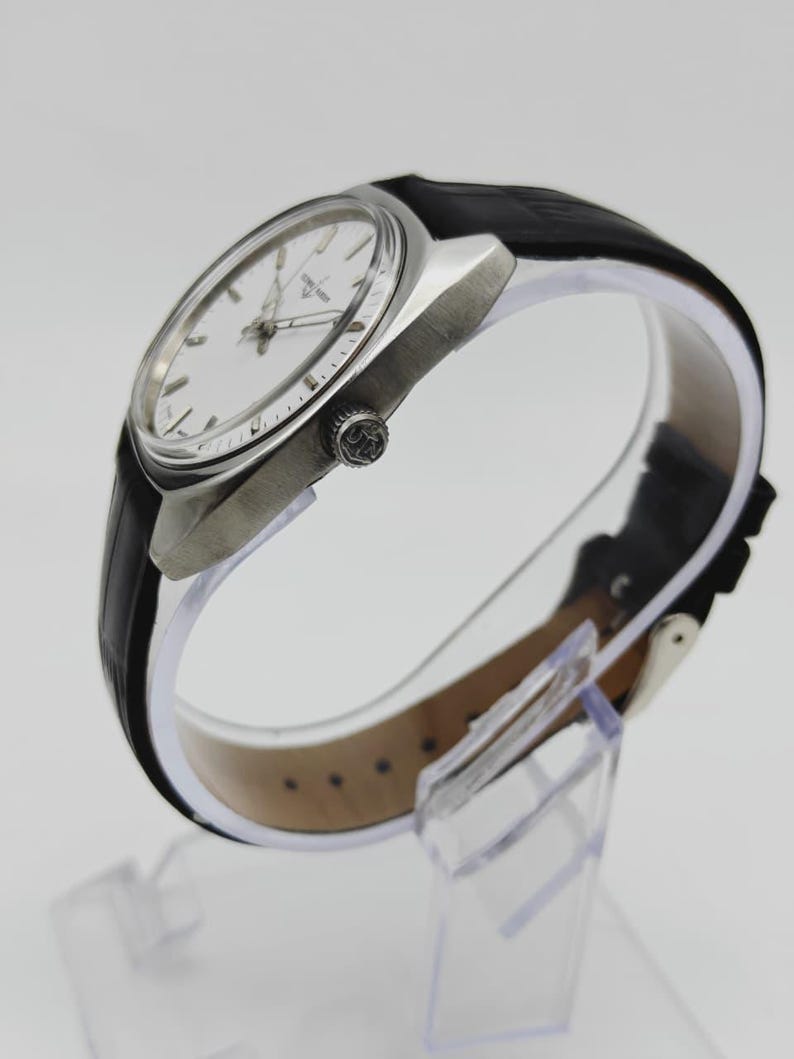 May include: A silver wristwatch with a white face and a black leather strap. The watch features a round face with silver hands and markers. The black strap is fastened with a silver buckle. The watch is displayed on a clear stand.