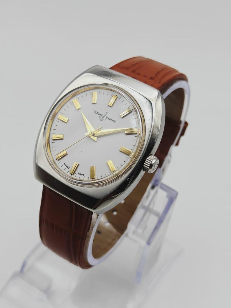 K&ouml;nnte beinhalten: Eine Vintage-Armbanduhr von Ulysse Nardin mit silberfarbenem Zifferblatt und goldfarbenen Stundenmarkierungen und Zeigern. Die Uhr hat ein Edelstahlgeh&auml;use und ein braunes Lederarmband. Die Uhr wird auf einem transparenten St&auml;nder pr&auml;sentiert.