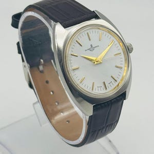 May include: A silver-toned wristwatch with a white face and gold hands and markers. The watch features the text "ULYSSE NARDIN" and a dark brown leather band.