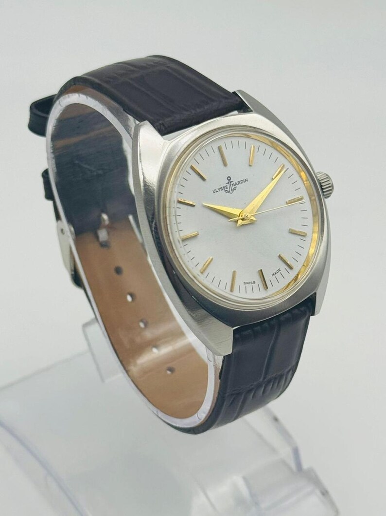 May include: A silver-toned wristwatch with a white face and gold hands and markers. The watch features the text "ULYSSE NARDIN" and a dark brown leather band.