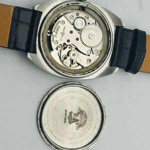May include: Vintage silver-toned watch with a black leather band. The watch face is open, revealing the intricate internal mechanisms, including gears and jewels. The back of the watch is detached, showing the brand name and details.