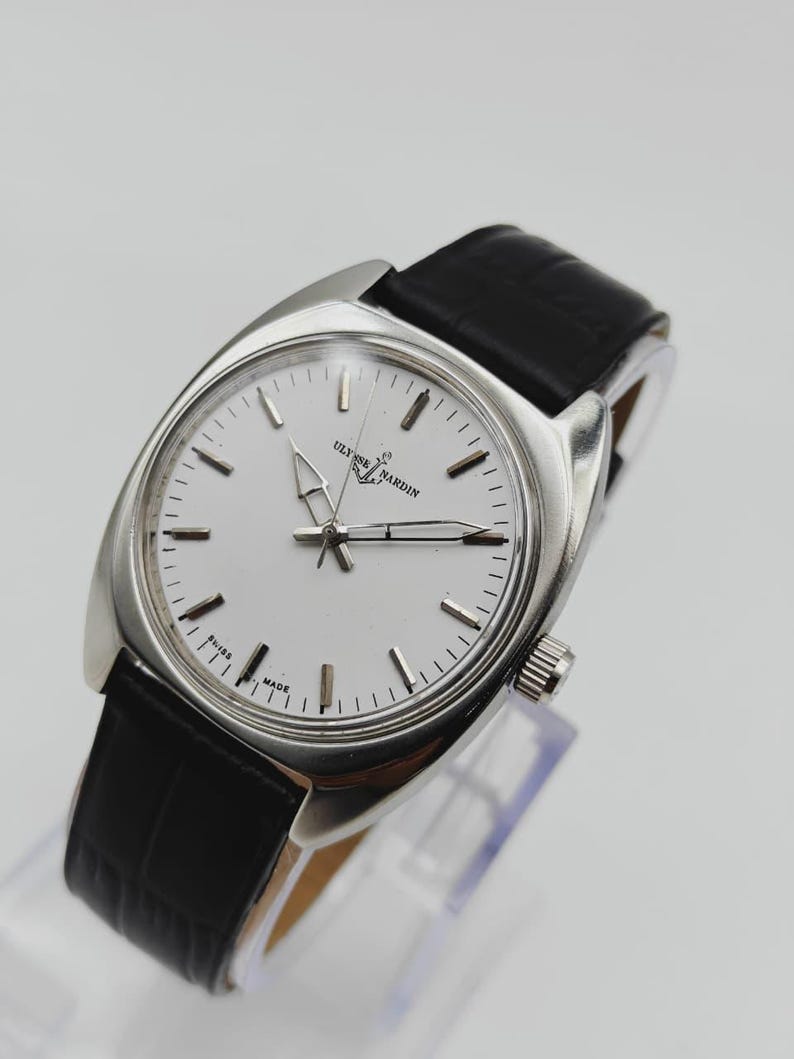 May include: A silver Ulysse Nardin wristwatch with a white face and black leather strap. The watch has a round face with silver hands and markers. The strap is black, and the watch is displayed on a clear stand.