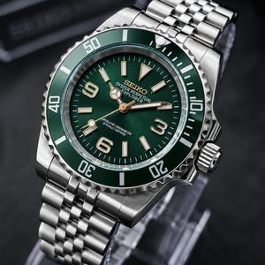 May include: A stainless steel wristwatch with a green dial and a matching green rotating bezel. The dial features the text SEIKO, OYSTER PERPETUAL, EXPLORER, and SUPERLATIVE CHRONOMETER OFFICIALLY CERTIFIED, with gold-tone hands and hour markers.