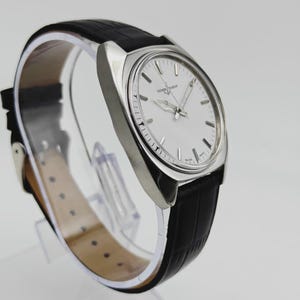 May include: A silver wristwatch with a white face and a black leather strap. The watch has silver hands and markers, and the strap has a textured pattern. The watch is displayed on a clear stand.
