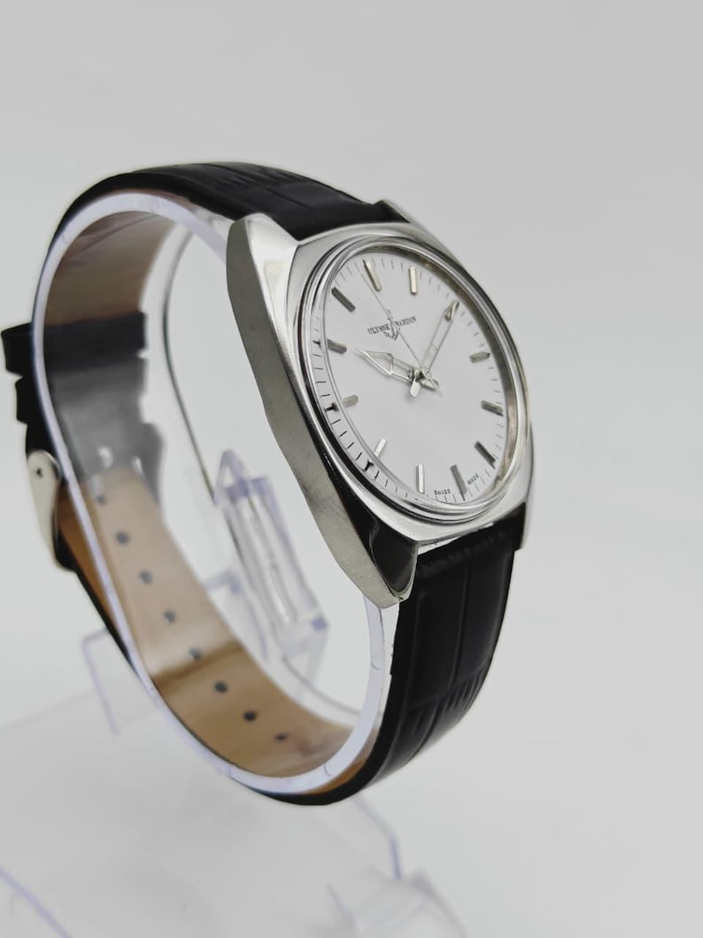 May include: A silver wristwatch with a white face and a black leather strap. The watch has silver hands and markers, and the strap has a textured pattern. The watch is displayed on a clear stand.