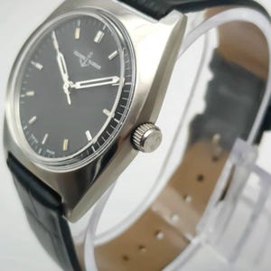 May include: A silver-toned wristwatch with a black face and a black leather band. The watch has a rectangular case and a silver crown. The watch face has silver hands and markers. The watch band is approximately 8 inches long.