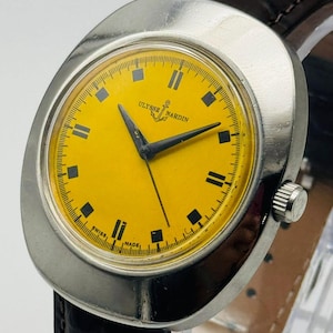 May include: A vintage Ulysse Nardin wristwatch with a bright yellow face and black hands. The watch has a silver-colored case and a dark brown leather strap. The watch face features black rectangular hour markers and the brand name.