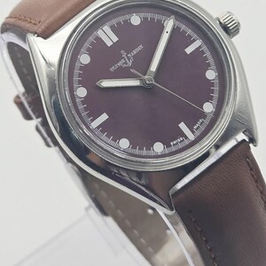 May include: A silver-toned wristwatch with a dark burgundy face and a brown leather band. The watch face features white hour markers and hands, with the brand name "Ulysse Nardin" displayed. The band is stitched and appears to be made of genuine leather.