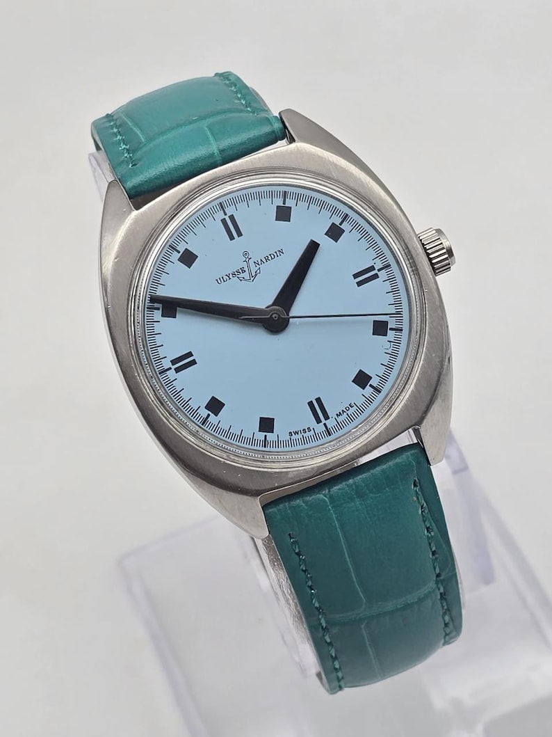Pu&ograve; includere: Un orologio da polso Ulysse Nardin argentato con quadrante azzurro e lancette nere. L'orologio ha un cinturino in pelle verde acqua. Il quadrante presenta indici neri rettangolari e le scritte "Ulysse Nardin" e "Swiss Made".