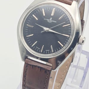 May include: A vintage Ulysse Nardin wristwatch with a black dial and silver-tone hands and markers. The watch has a stainless steel case and a brown leather strap. The watch is displayed on a clear stand.