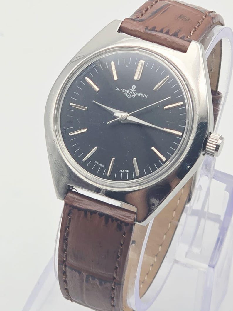 May include: A vintage Ulysse Nardin wristwatch with a black dial and silver-tone hands and markers. The watch has a stainless steel case and a brown leather strap. The watch is displayed on a clear stand.