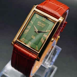Puede incluir: Un reloj Seiko Quartz dorado con una esfera rectangular verde y n&uacute;meros romanos dorados. El reloj tiene una correa de cuero roja. La esfera del reloj presenta el logotipo de Seiko y un patr&oacute;n texturizado. El reloj se exhibe sobre un soporte transparente.