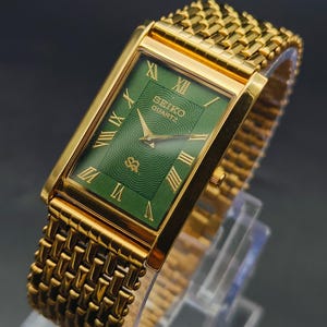 Puede incluir: Reloj Seiko Quartz dorado con esfera verde rectangular y números romanos dorados. El reloj tiene una correa dorada tejida. La esfera del reloj presenta el logotipo de Seiko Quartz y un patrón decorativo.