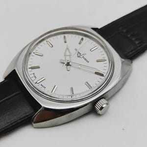 May include: A silver wristwatch with a white face and a black leather strap. The watch face features silver hour markers and hands. The brand name "ULYSSE NARDIN" is printed on the watch face. The watch has a classic, elegant design.