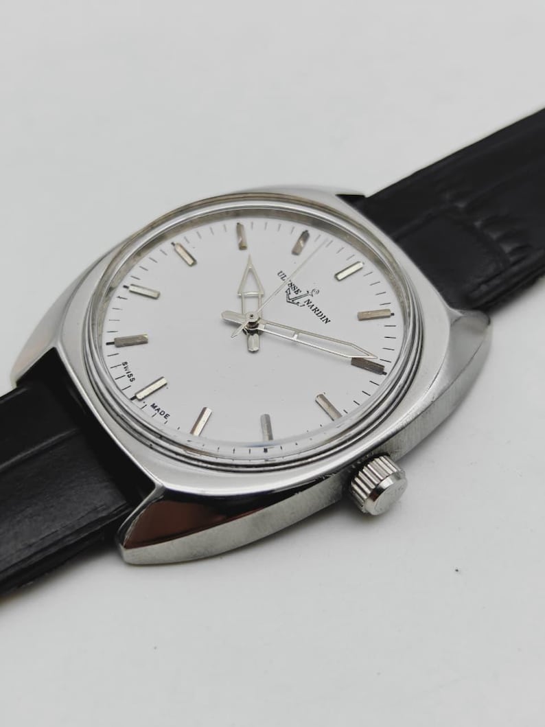 May include: A silver wristwatch with a white face and a black leather strap. The watch face features silver hour markers and hands. The brand name "ULYSSE NARDIN" is printed on the watch face. The watch has a classic, elegant design.