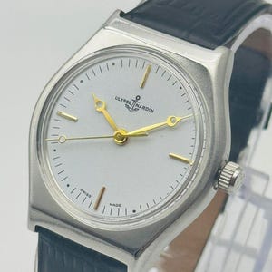 May include: A silver-toned Ulysse Nardin wristwatch with a white face, gold hands, and a black leather band. The watch face features the brand name and the words "Swiss Made". The watch has a rectangular case and a crown on the side.