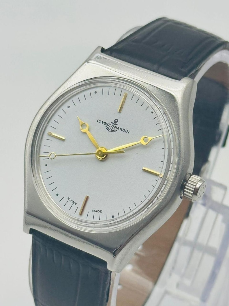 May include: A silver-toned Ulysse Nardin wristwatch with a white face, gold hands, and a black leather band. The watch face features the brand name and the words "Swiss Made". The watch has a rectangular case and a crown on the side.