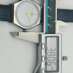 May include: A silver-toned wristwatch with a light-colored face and gold hands is measured with a digital caliper. The watch has a black leather band. The caliper displays a measurement of 38.06 mm.
