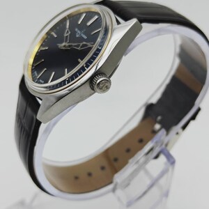 Puede incluir: Un reloj de pulsera cl&aacute;sico con esfera negra y caja plateada. El reloj presenta agujas e indicadores dorados, y una correa de cuero negro. El reloj se exhibe sobre un soporte transparente.