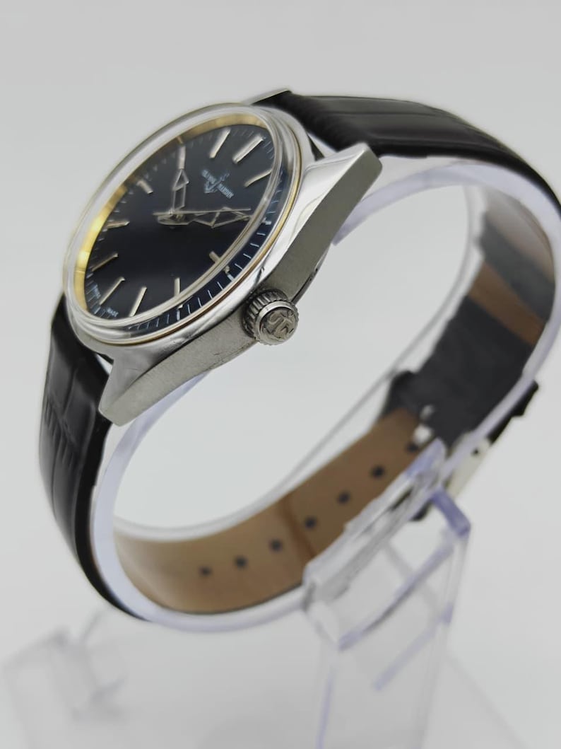 Puede incluir: Un reloj de pulsera cl&aacute;sico con esfera negra y caja plateada. El reloj presenta agujas e indicadores dorados, y una correa de cuero negro. El reloj se exhibe sobre un soporte transparente.