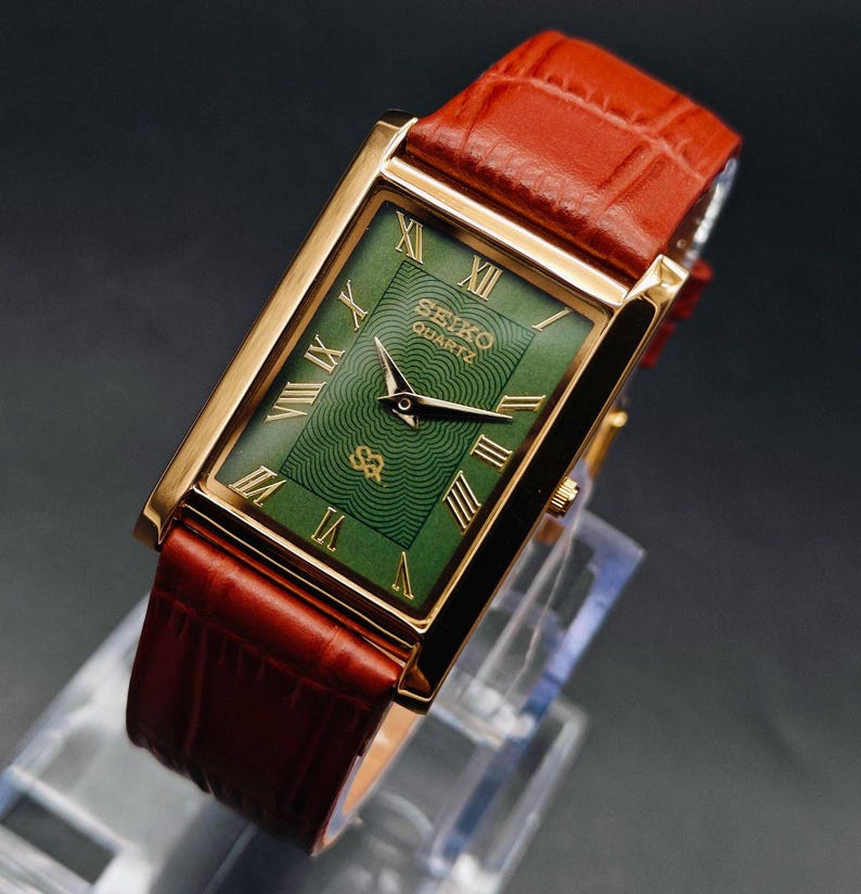 Puede incluir: Un reloj Seiko Quartz rectangular con caja dorada y esfera verde con n&uacute;meros romanos. El reloj tiene una correa de cuero marr&oacute;n. El reloj se exhibe sobre un soporte transparente.