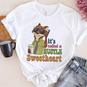 May include: White t-shirt with a cartoon fox wearing sunglasses, a green shirt, and a tie, holding a coffee cup. The shirt has the text "it's called a HUSTLE Sweetheart" in colourful retro lettering.