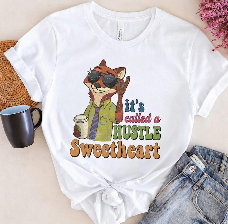 May include: White t-shirt with a cartoon fox wearing sunglasses, a green shirt, and a tie, holding a coffee cup. The shirt has the text "it's called a HUSTLE Sweetheart" in colourful retro lettering.