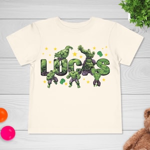 May include: Cream-colored t-shirt featuring the name "LUCAS" in large, green, comic-style letters. The letters are decorated with images of a green superhero. Small yellow stars are scattered around the name and figures.