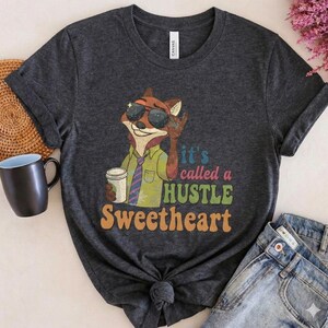 May include: Dark grey t-shirt with a cartoon fox wearing sunglasses and holding a coffee cup. The shirt features the text "It's called a HUSTLE Sweetheart" in retro lettering. The fox is wearing a green shirt and a blue and purple tie.