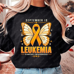 Könnte beinhalten: Ein schwarzes Sweatshirt mit einem grafischen Design und dem Text SEPTEMBER IS LEUKEMIA CANCER AWARENESS. Das Motiv zeigt eine orangefarbene Schleife, die zwischen zwei Schmetterlingsflügeln in Orange-, Gelb- und Weißtönen zentriert ist.