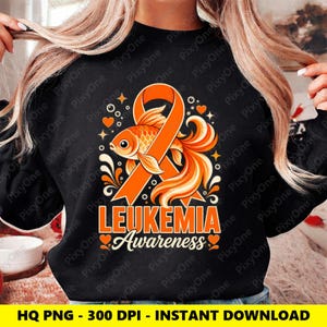 Puede incluir: Una sudadera negra de manga larga con un gráfico de un lazo de concienciación naranja entrelazado con un pez dorado. El diseño incluye el texto naranja LEUKEMIA Awareness, rodeado de pequeños corazones naranjas y remolinos decorativos.