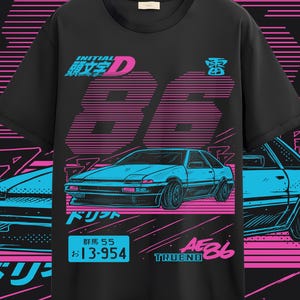 Puede incluir: Camiseta negra con un gráfico turquesa y rosa de un coche, texto japonés y el número 86. El diseño incluye las palabras "INITIAL D", "TRUENO" y "AE86". El gráfico del coche es una ilustración estilizada.