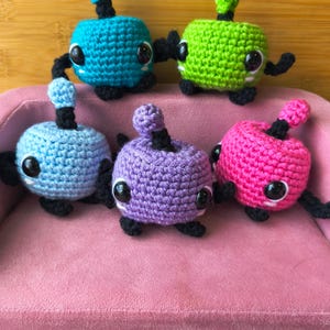 May include: A collection of five colorful, crocheted creatures. Each has a round body, black arms and legs, and a black antenna-like feature. The creatures are in shades of blue, green, purple, and pink, with large, round eyes.